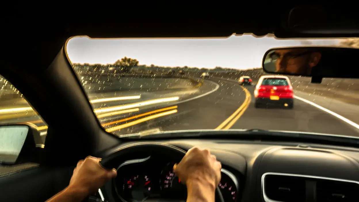 Vista interior de um carro a conduzir numa estrada curva com dois carros à frente e mãos no volante.