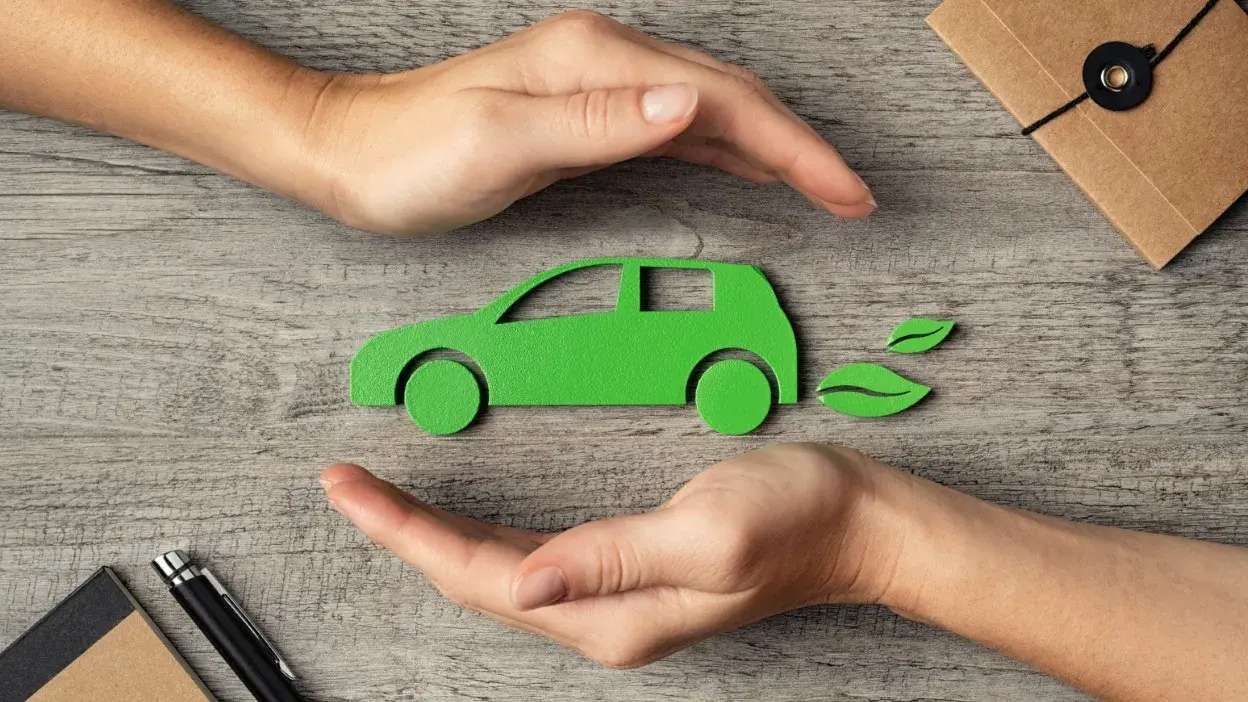 Mãos a proteger um carro verde de papel com folhas a sair do escape, simbolizando sustentabilidade ambiental.
