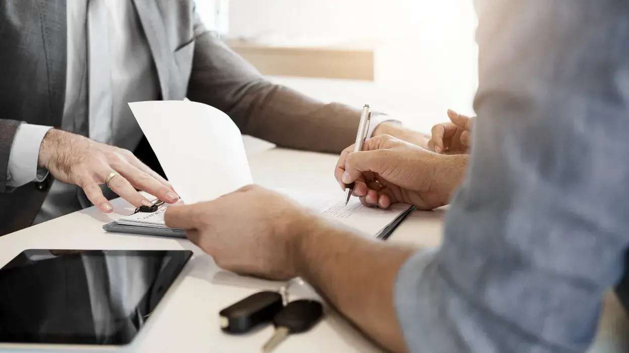 Duas pessoas a assinar documentos numa mesa com chaves de carro e um tablet visíveis.