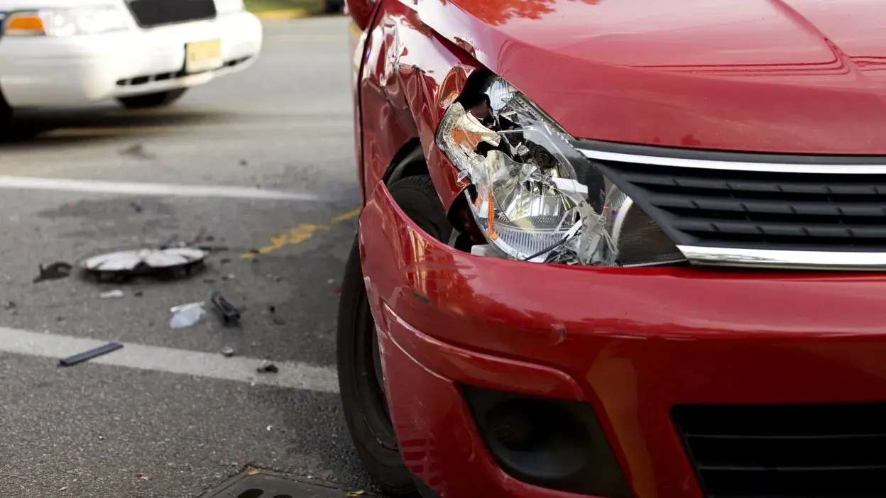 Carro vermelho com o farol e para-choques danificados após colisão, com detritos no chão.