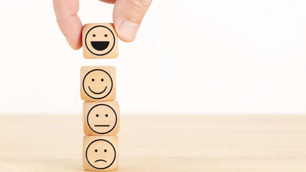 Cubos de madeira empilhados com rostos sorridente, neutro e triste, mão a colocar cubo sorridente no topo