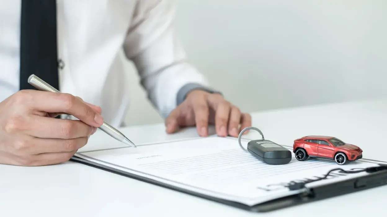 Pessoa a assinar documento com chave e miniatura de carro sobre a mesa