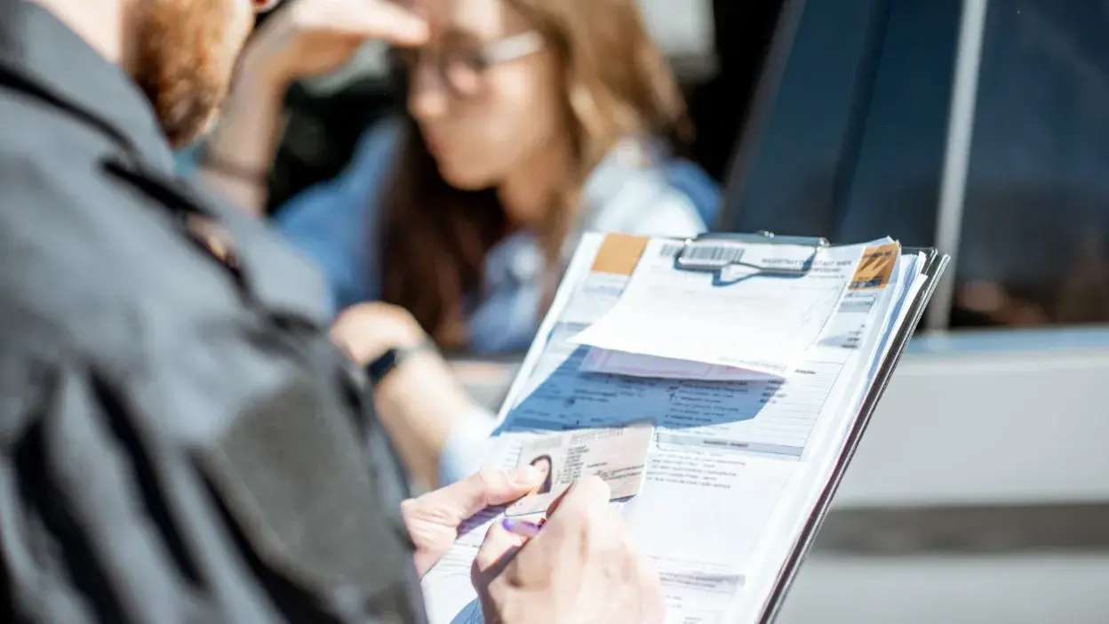 Agente da autoridade a verificar documentos de condutor junto a viatura, com mulher ao fundo.