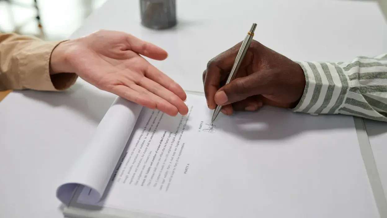 Mão a assinar documento enquanto outra mão aponta para a assinatura.