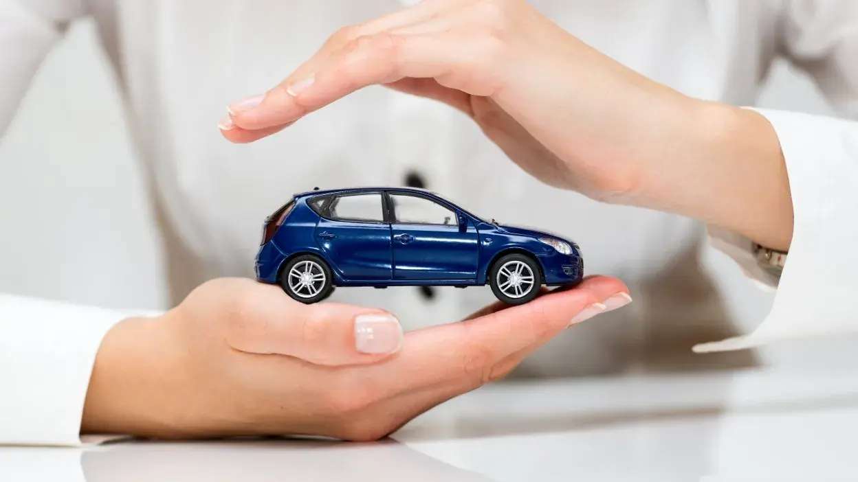 Mãos a proteger um carro em miniatura azul sobre uma superfície branca.