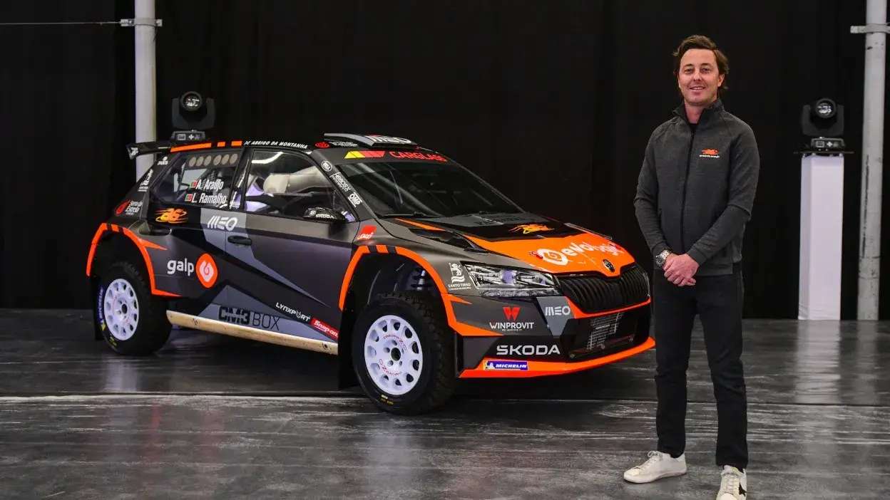 Piloto Armindo Araújo em pé ao lado do seu carro de rali preto e laranja numa garagem.