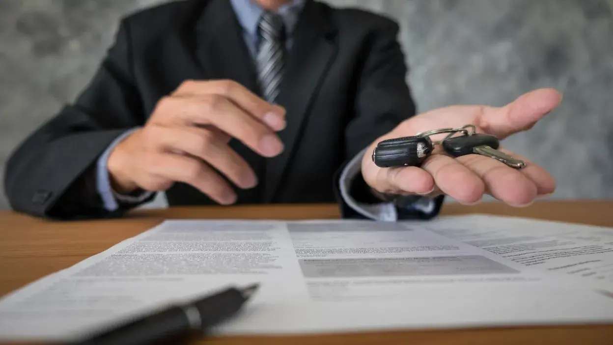 Pessoa de fato a entregar chaves de carro sobre mesa com documentos e caneta.