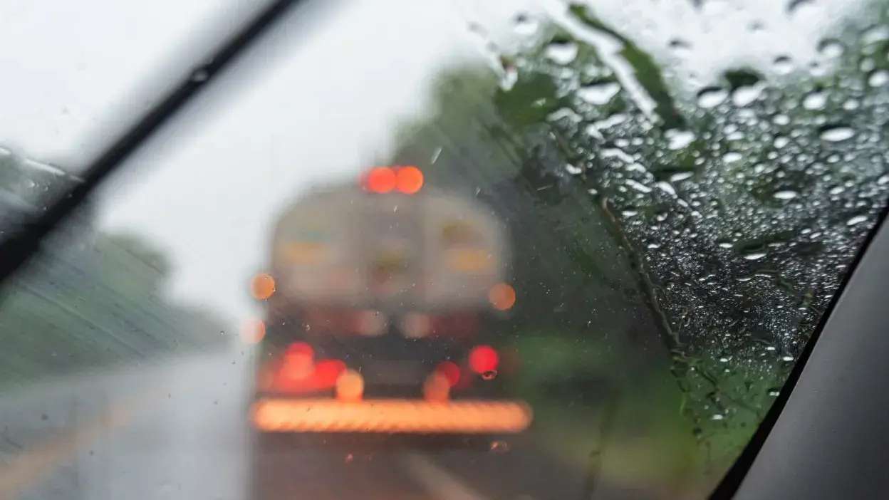 Vista através do vidro de um carro com gotas de chuva e limpa-vidros em funcionamento, com veículo à frente desfocado na estrada molhada.