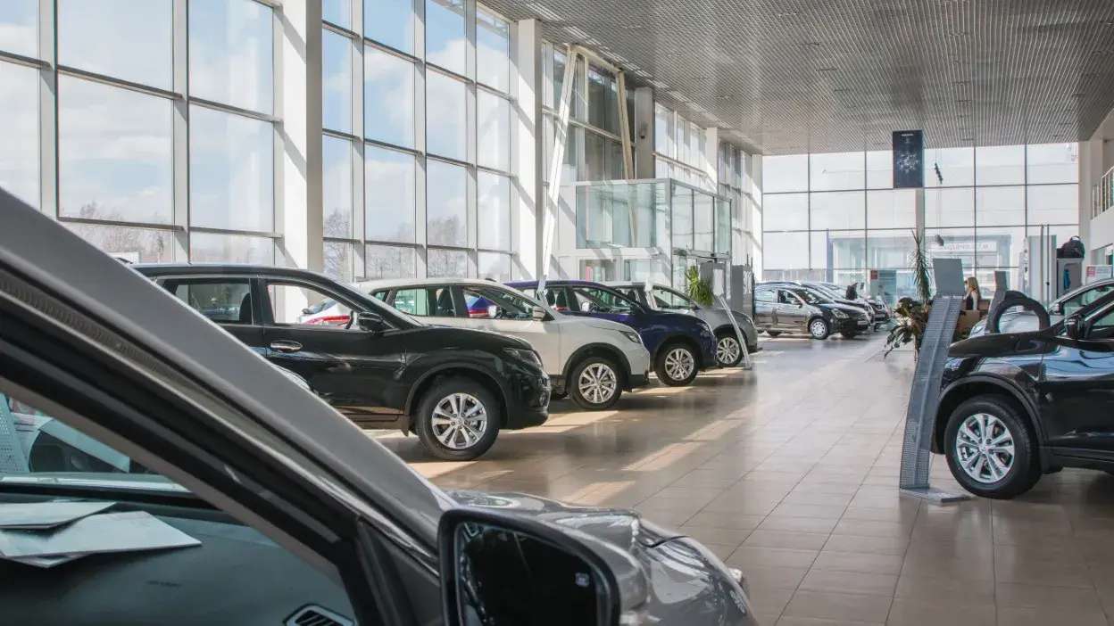 Interior de concessionário com vários carros usados expostos em fila, com grande iluminação natural.