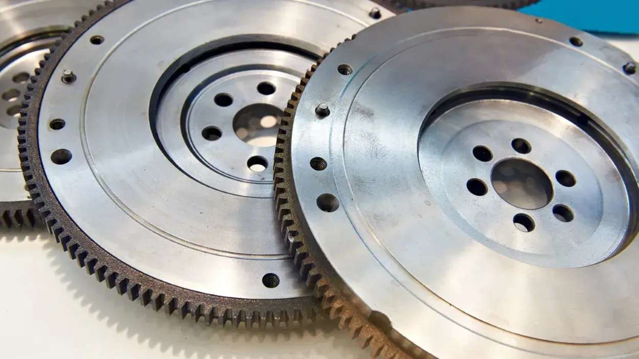 Dois volantes bimassa metálicos com dentes e furos circulares, sobre superfície branca.