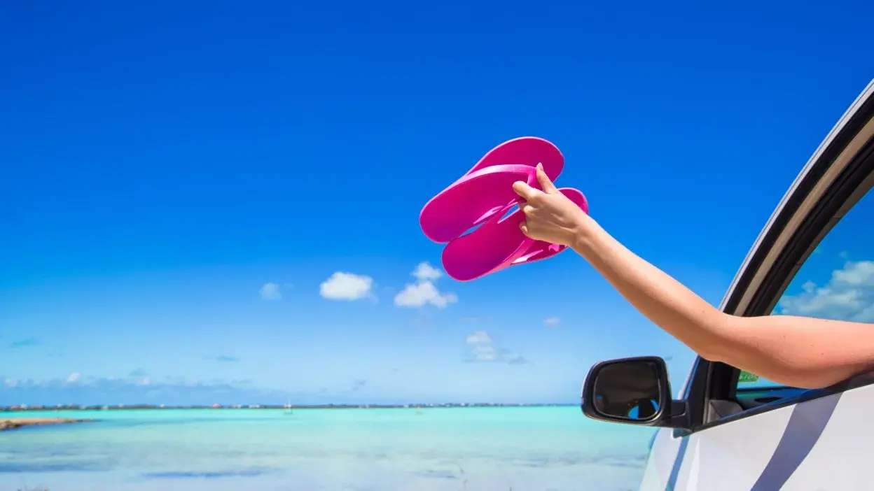 Braço de pessoa a segurar chinelos cor-de-rosa fora da janela de um carro junto ao mar e céu azul.