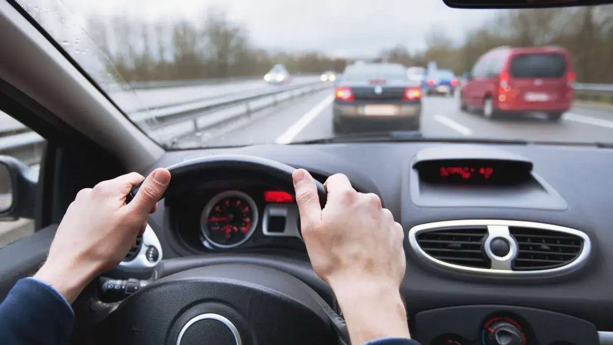 Vista do interior de um carro com as mãos no volante, a conduzir numa estrada com trânsito à frente.