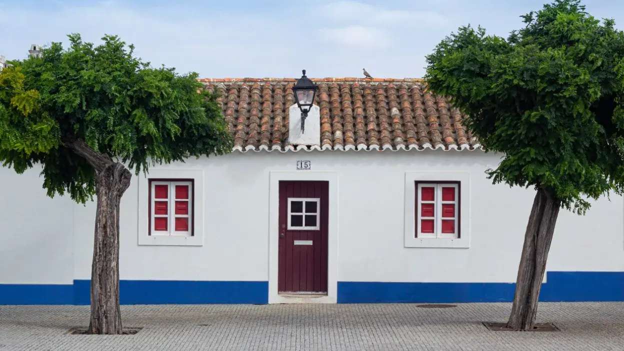 Fachada branca com porta e janelas vermelhas, telhado de telha e duas árvores em frente