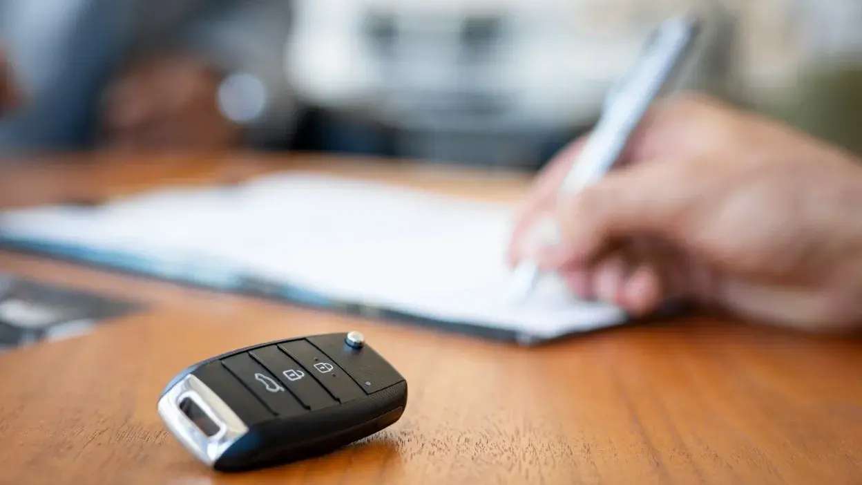 Chave de carro sobre mesa com pessoa a assinar documento ao fundo