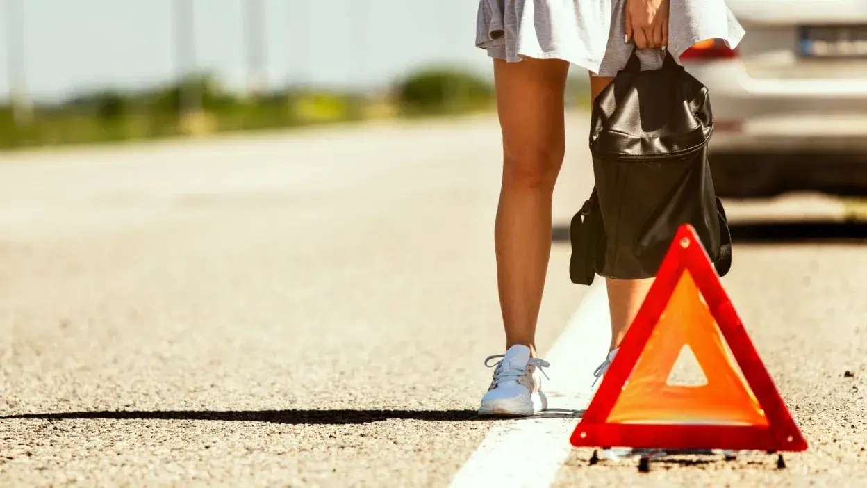Pessoa de pé junto a um triângulo de sinalização refletor na berma da estrada, com uma mochila preta na mão e um carro estacionado ao fundo.
