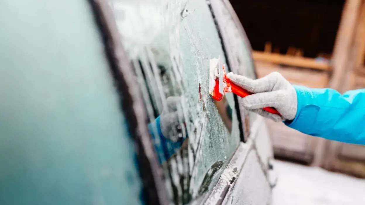 Pessoa a remover gelo do vidro lateral de um carro com um raspador vermelho, usando luvas e casaco azul.