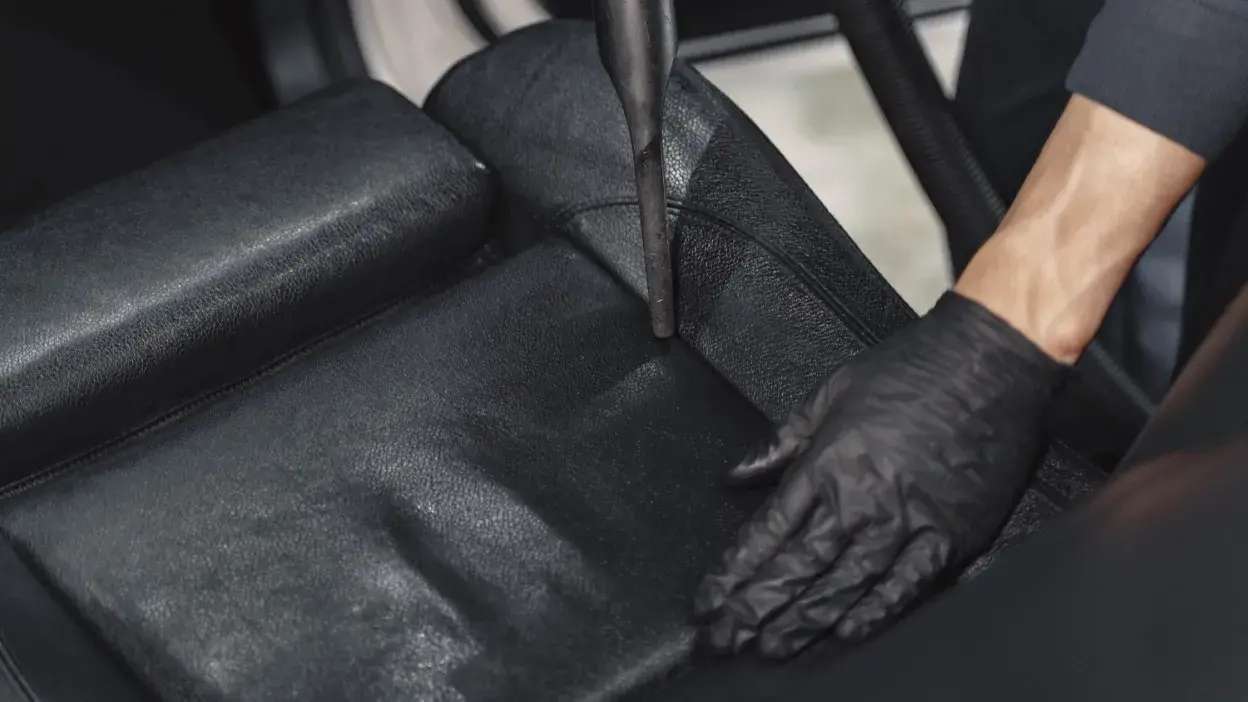 Pessoa a limpar estofos de couro preto de um carro com equipamento profissional e luvas pretas.