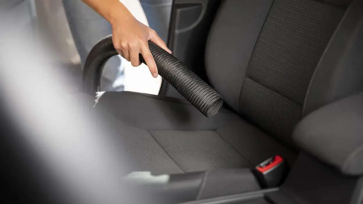 Pessoa a aspirar o banco de um carro com um tubo de aspirador preto.