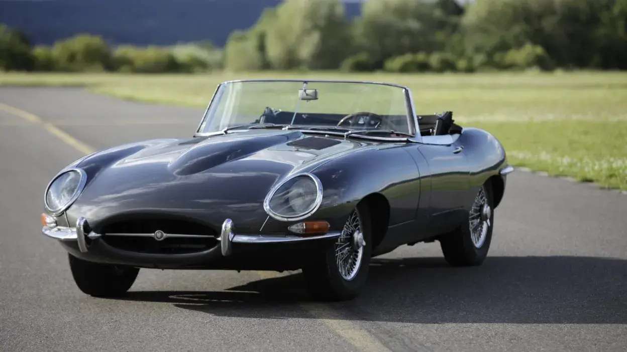 Carro clássico descapotável preto estacionado numa estrada com relva ao fundo.