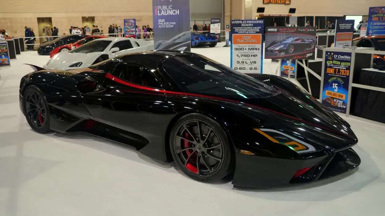 Carro desportivo preto com faixa vermelha exposto numa feira automóvel com outros carros ao fundo.