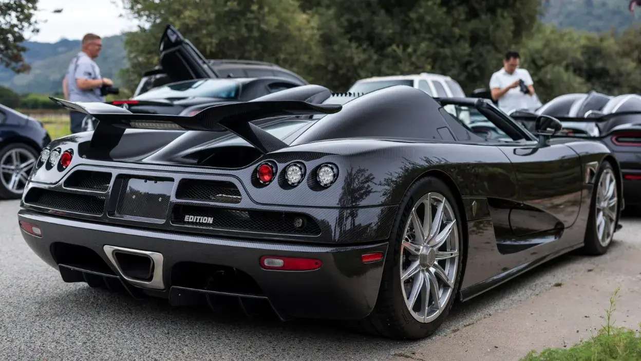Vista traseira de um carro desportivo preto com spoiler, estacionado numa estrada com pessoas ao fundo.