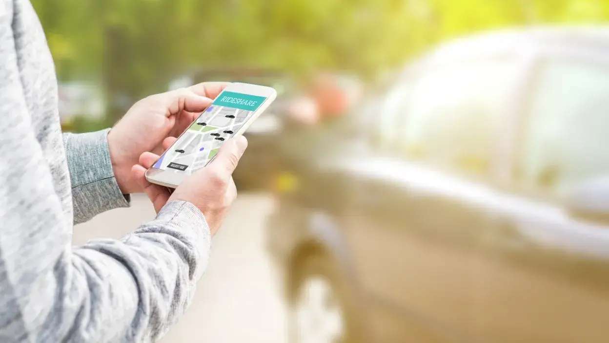 Pessoa a usar aplicação de carpooling num smartphone junto a um carro estacionado.