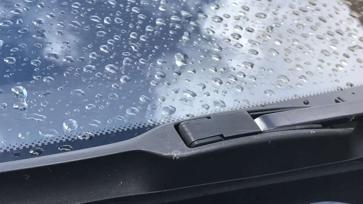 Limpa para-brisas de carro com gotas de água no vidro e céu refletido.