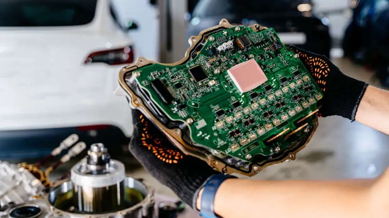 Mãos com luvas a segurar uma centralina eletrónica de carro, com componentes eletrónicos visíveis.