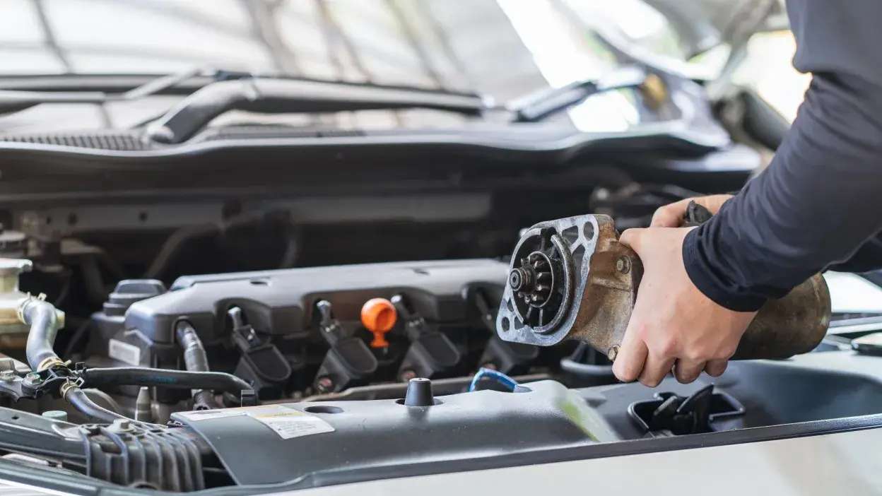 Mãos a segurar um motor de arranque junto ao motor de um carro aberto