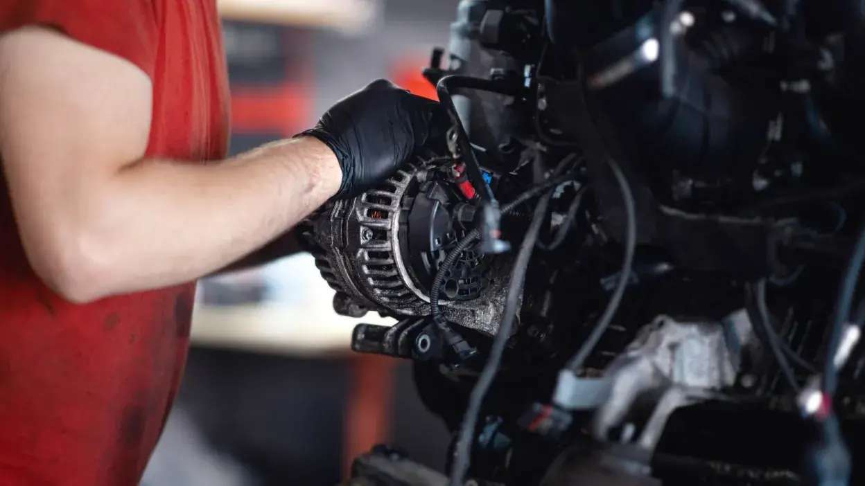 Mão de mecânico com luva preta a trabalhar num alternador de carro no motor.