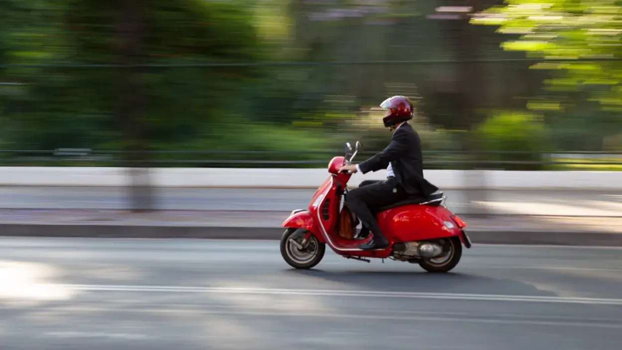Homem de fato e capacete vermelho a conduzir um ciclomotor vermelho numa estrada urbana
