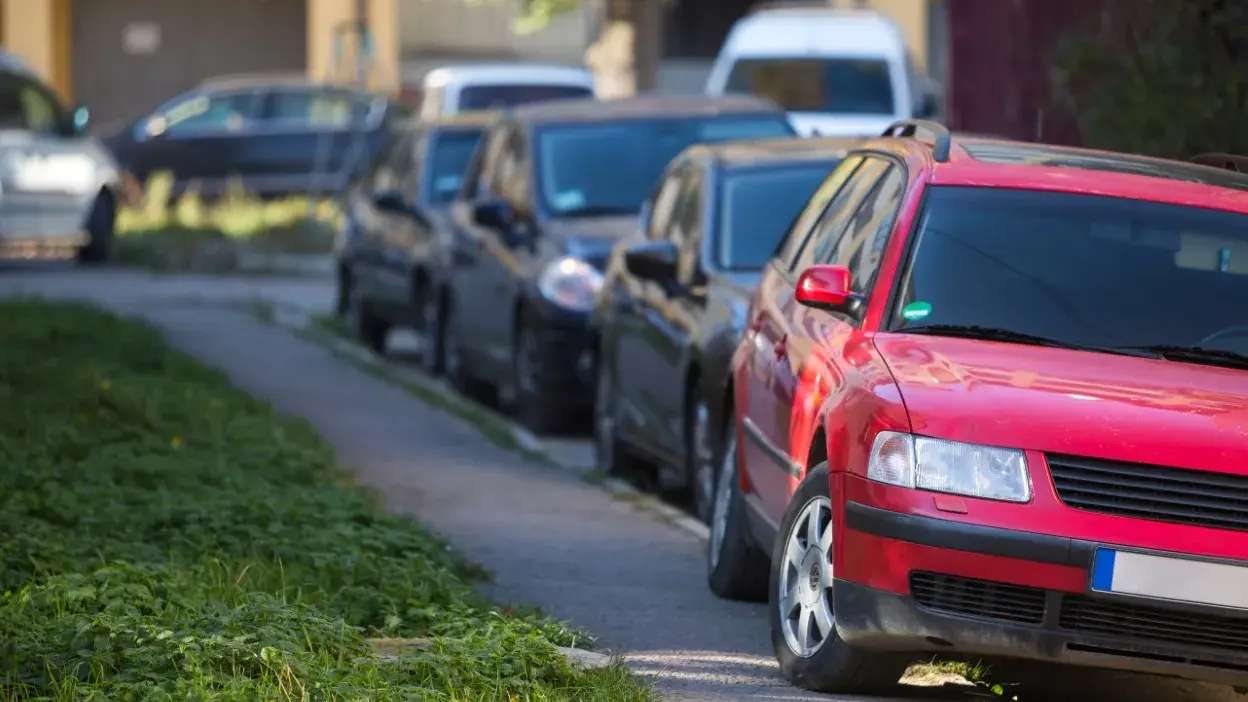 Veículo vermelho estacionado parcialmente em cima do passeio numa rua com outros carros estacionados em fila.