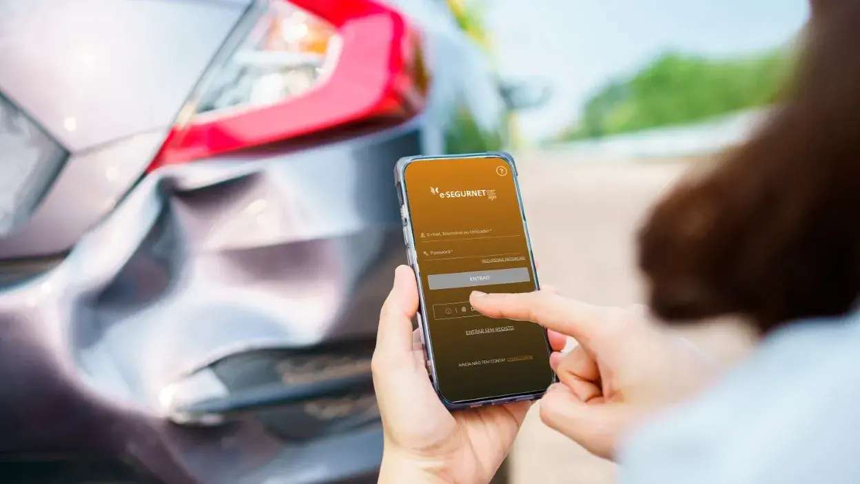 Pessoa a usar aplicação móvel para declaração amigável após acidente automóvel com carro danificado ao fundo