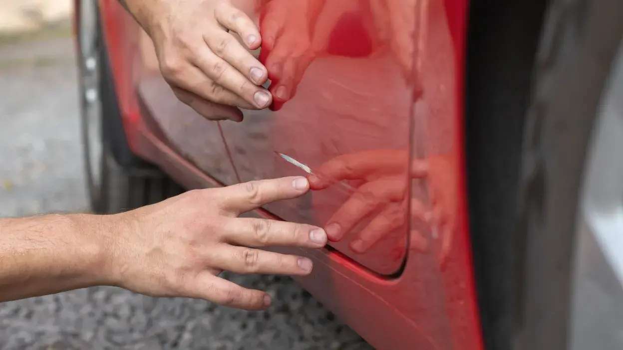Mãos a inspecionar um risco na porta de um carro vermelho