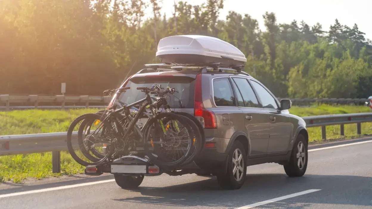 Carro cinzento com bicicletas na bagageira traseira e mala de tejadilho a circular numa estrada rodeada de árvores.