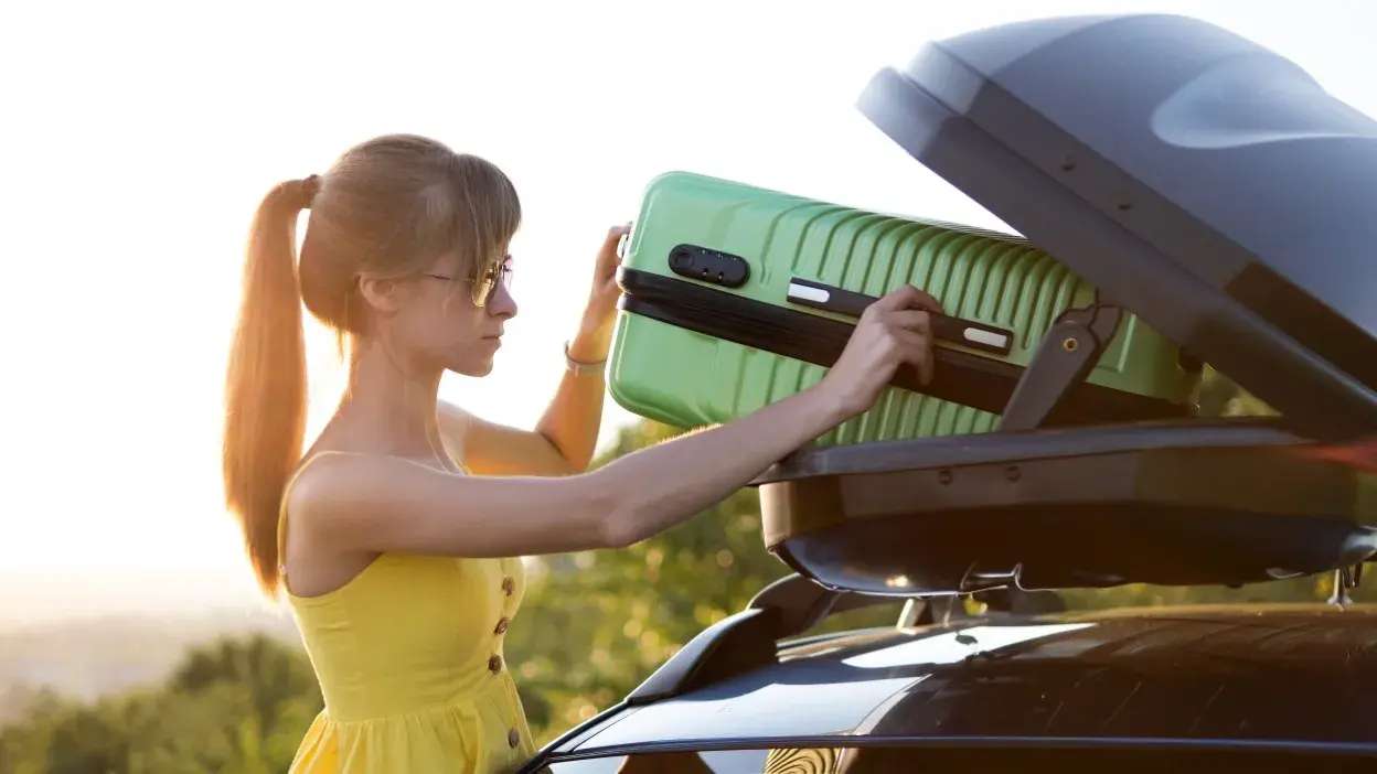 Mulher a colocar mala verde numa caixa de bagagem no tejadilho de um carro.