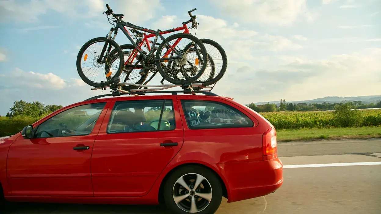 Carro vermelho em movimento com duas bicicletas presas no tejadilho numa estrada rural.