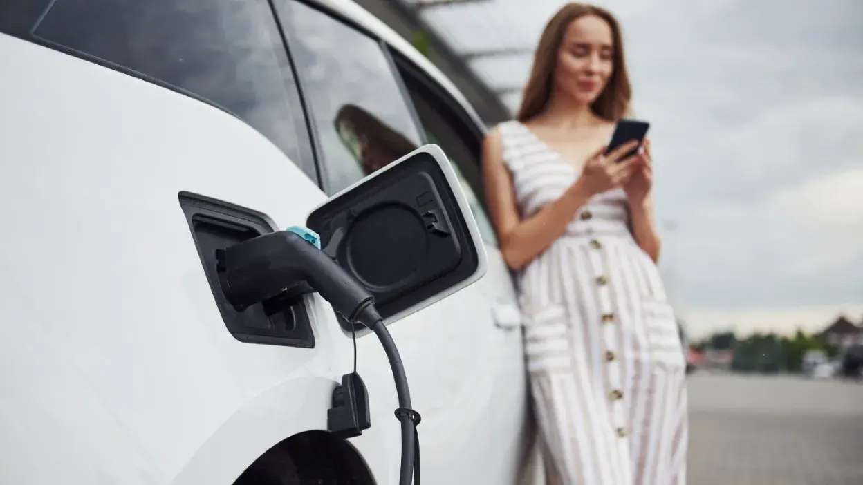 Carro elétrico branco a ser carregado com mulher ao lado a usar telemóvel.