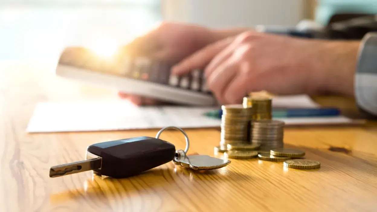 Chave de carro e moedas sobre mesa com pessoa a usar calculadora ao fundo