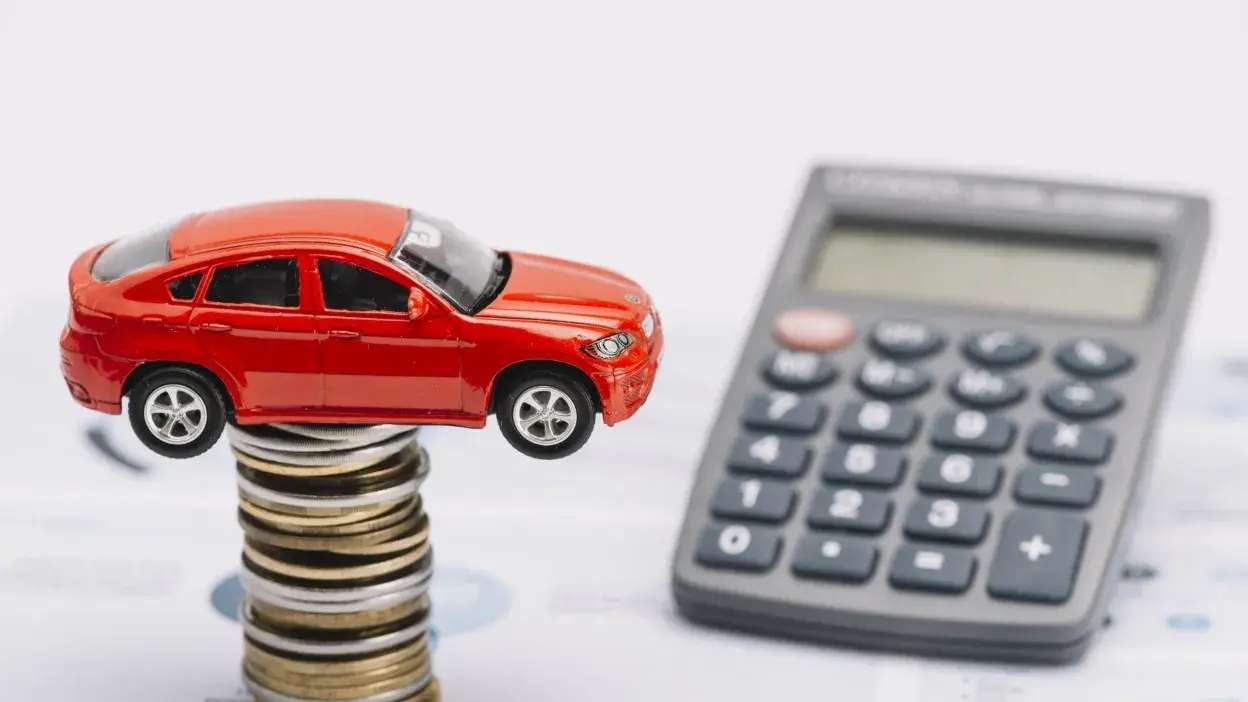 Carro vermelho em miniatura sobre pilha de moedas com calculadora ao fundo.