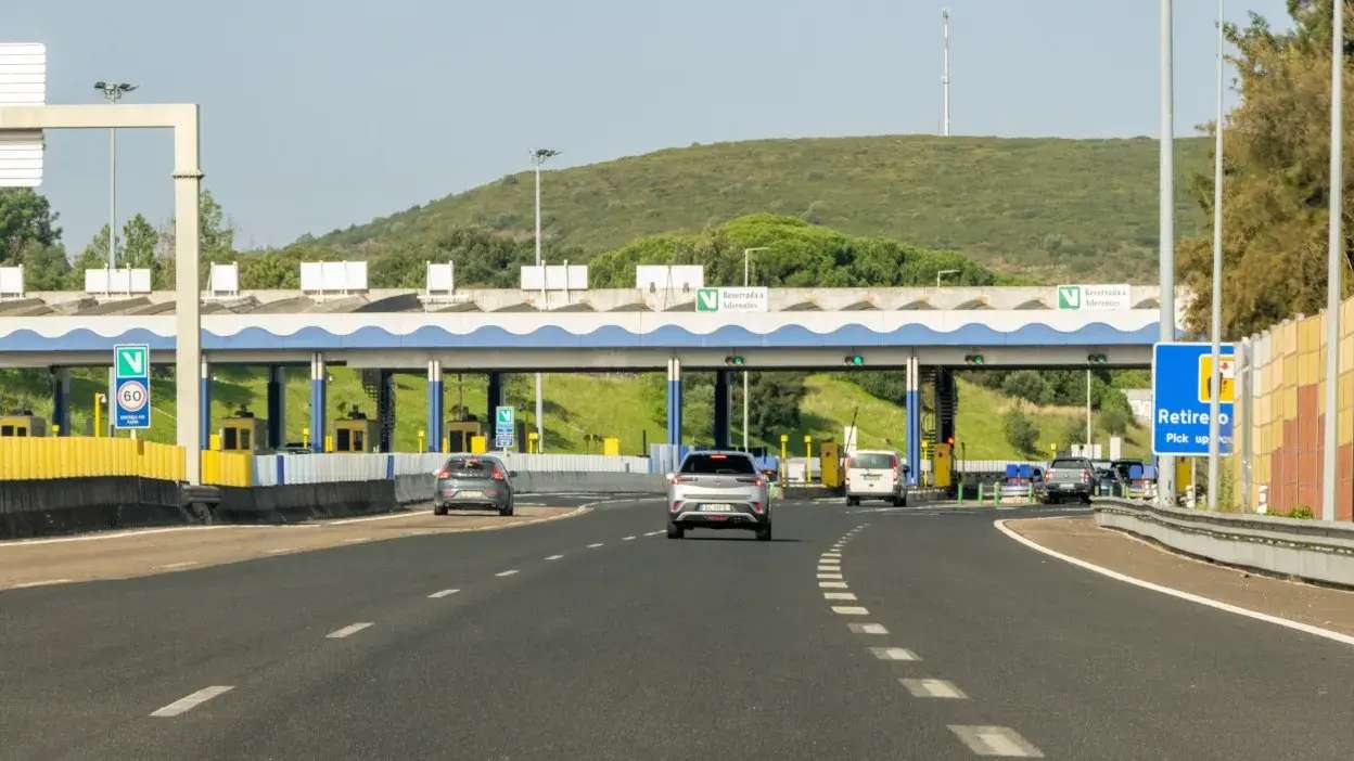 Viaturas a aproximarem-se de uma portagem numa autoestrada com sinalização visível e colinas ao fundo