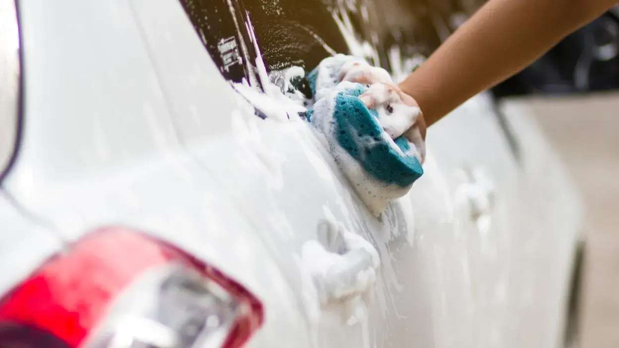 Mão a lavar carro branco com esponja e espuma.