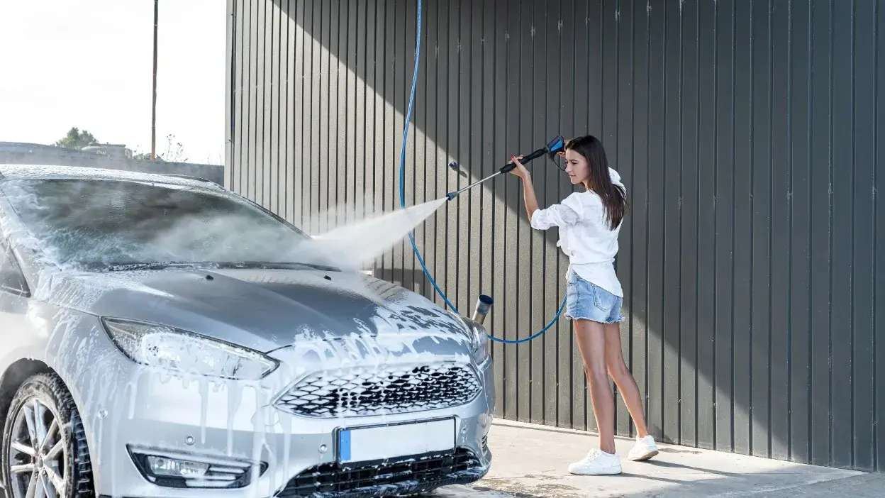 Mulher a lavar um carro prateado com máquina de pressão numa área exterior.