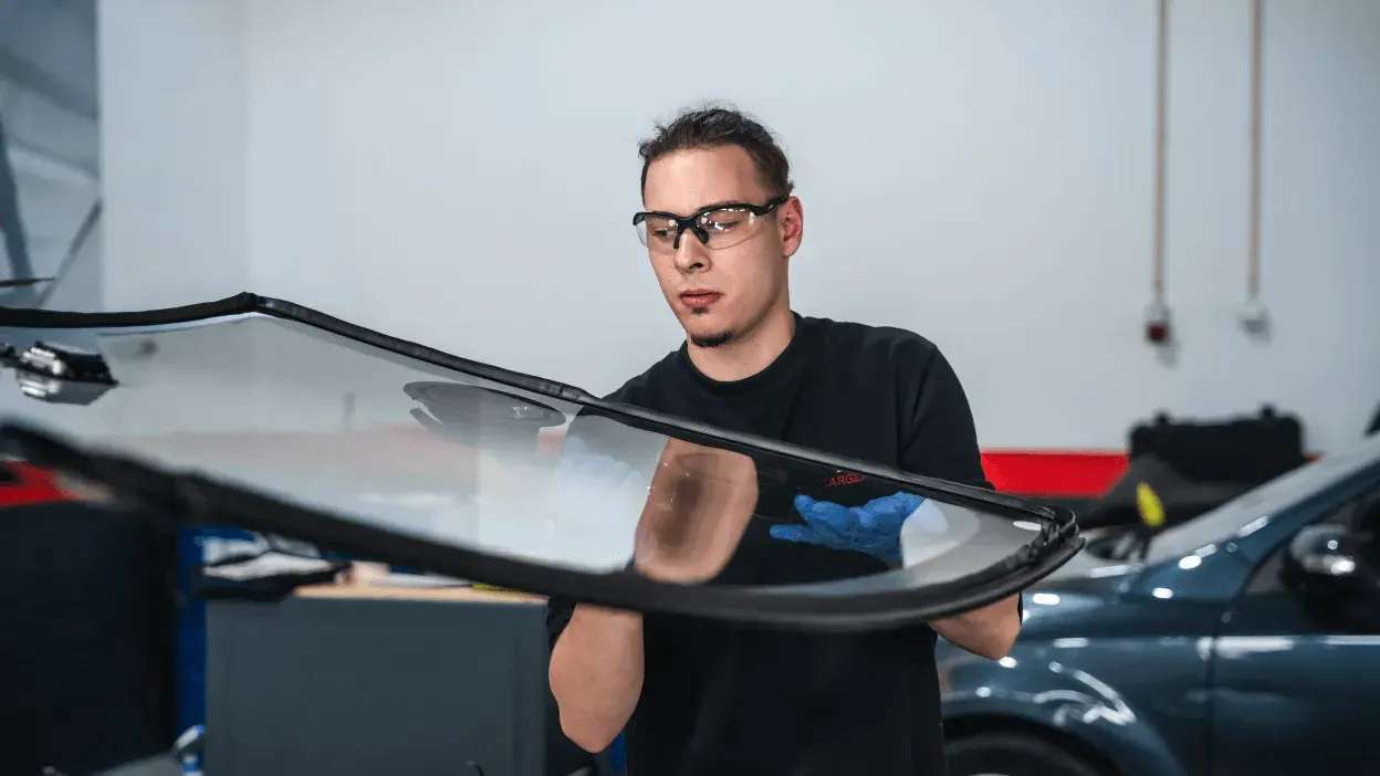 Homem com luvas e óculos de proteção a manusear um vidro de carro numa oficina automóvel