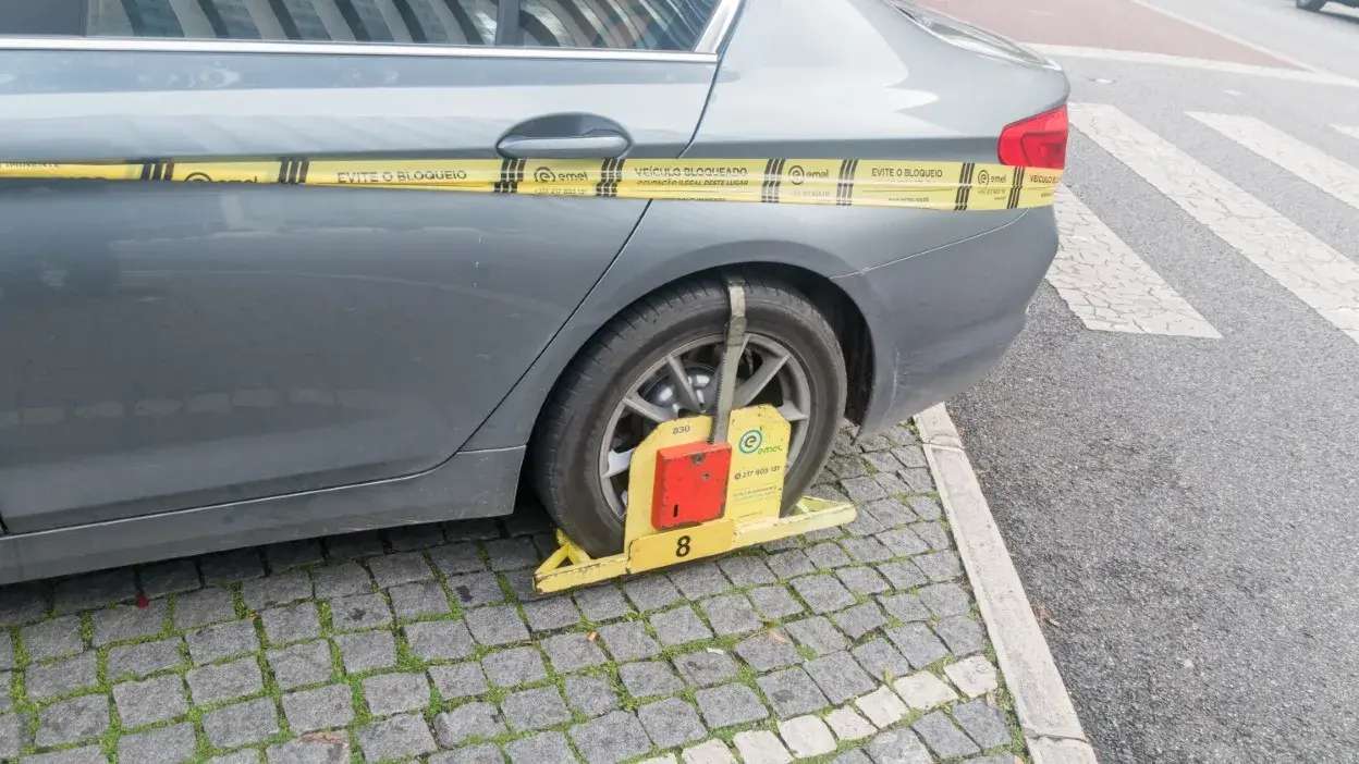 Veículo cinzento com roda traseira bloqueada por dispositivo da EMEL numa rua com calçada e faixa de peões.
