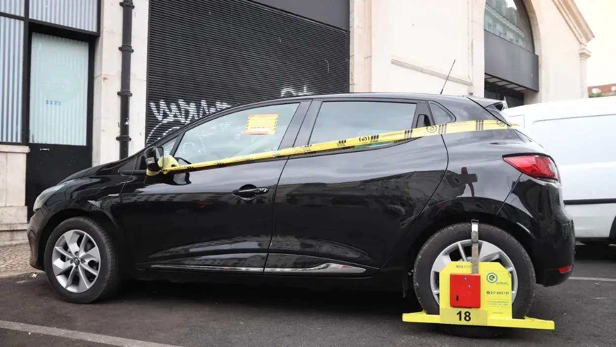 Carro preto com roda traseira bloqueada pela EMEL e fita amarela à volta do veículo estacionado na rua