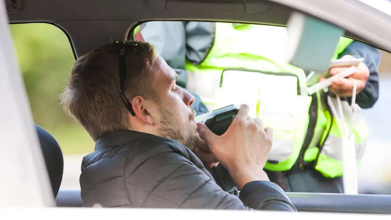 Homem a fazer teste de alcoolemia dentro de carro com polícia ao lado.