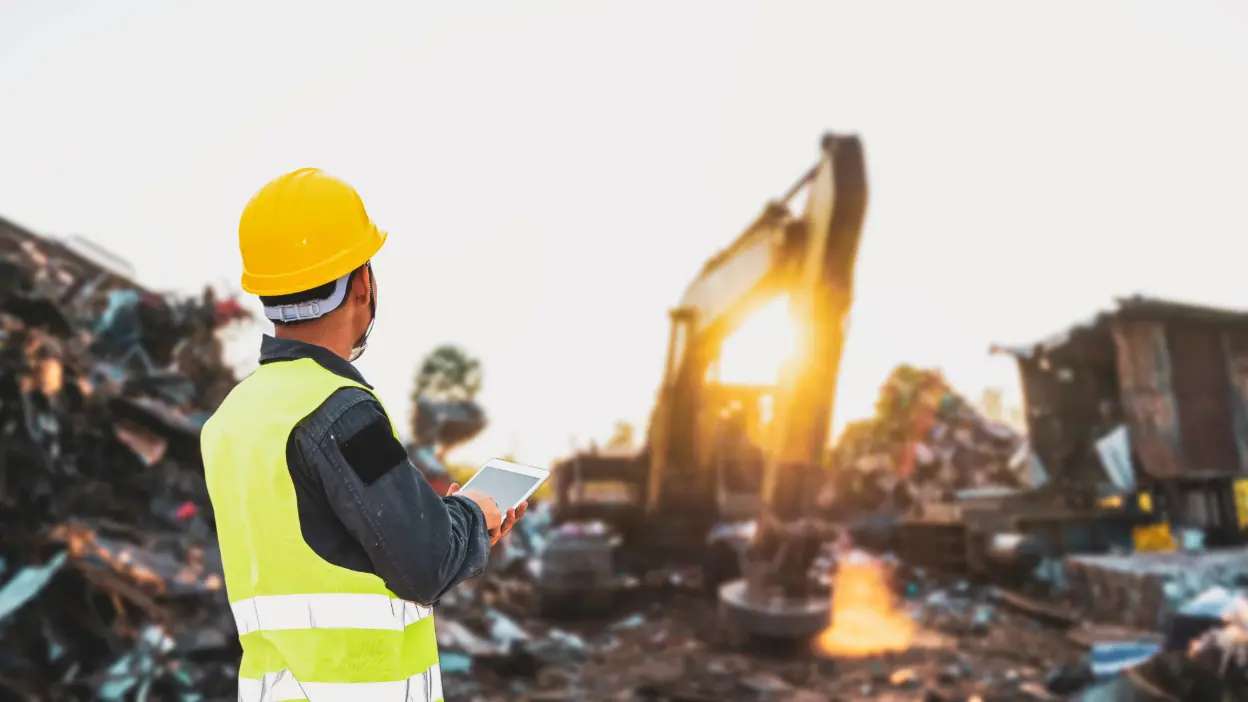 Trabalhador com capacete e colete refletor a usar tablet numa área de demolição com maquinaria pesada ao fundo
