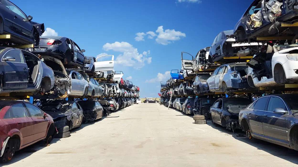 Pátio com várias filas de carros empilhados e danificados para abate, sob céu azul com nuvens.