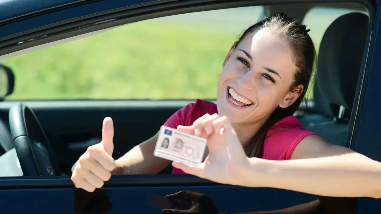 Mulher sorridente dentro de carro a mostrar carta de condução e a fazer sinal de positivo com a mão.